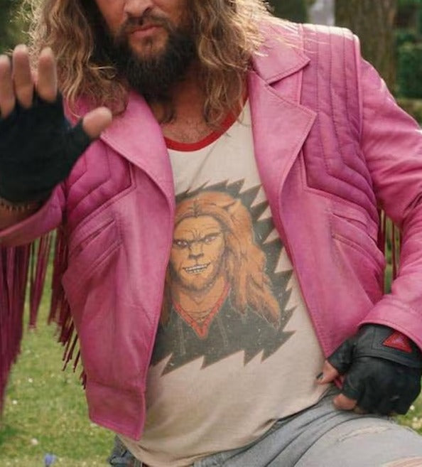 A man wearing a pink leather jacket with fringe over a graphic t-shirt featuring a cartoon character, set outdoors on grass.