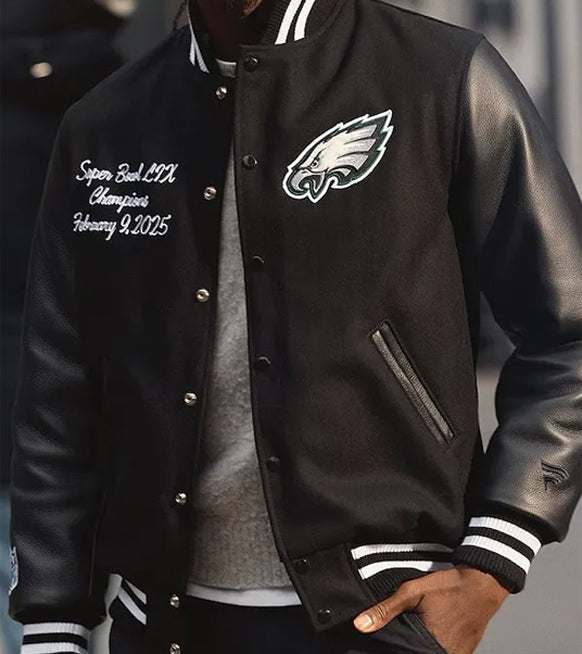 A close-up of a black varsity-style jacket worn by a person. The jacket has white and black striped ribbed cuffs and collar, a front button closure, a zip pocket, and embroidered Philadelphia Eagles branding and championship text on the chest.