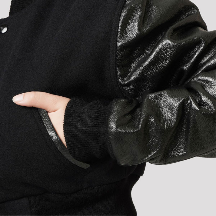 Close-up of a black knit jacket with glossy black faux-leather sleeves, showing a hand in a pocket against a light gray background.