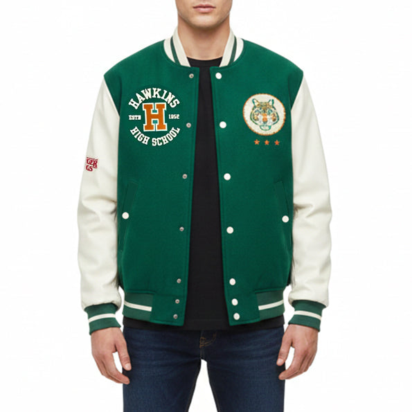 A man wearing a green and cream varsity jacket with 'Hawkins High School' embroidery on the left chest and a tiger crest on the right chest, paired with a black t-shirt and blue jeans against a white background.
