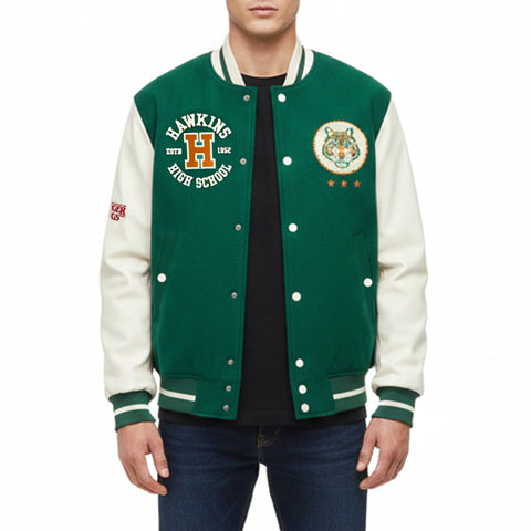 A man wearing a green and cream varsity jacket with 'Hawkins High School' embroidery on the left chest and a tiger crest on the right chest, paired with a black t-shirt and blue jeans against a white background.